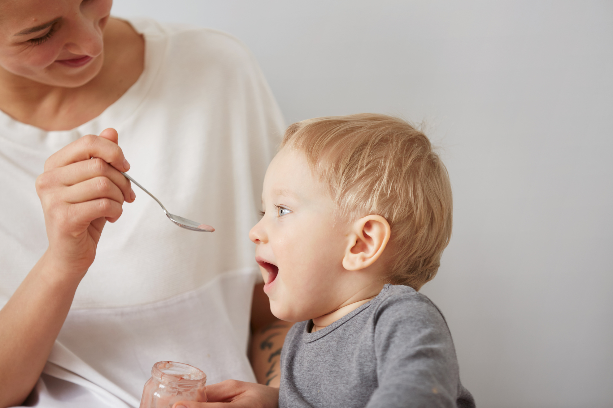 Young attractive mother with her one years old little son dressed in pajamas  giving him his first fruit smoothies food in the bedroom at the weekend together, lazy morning.